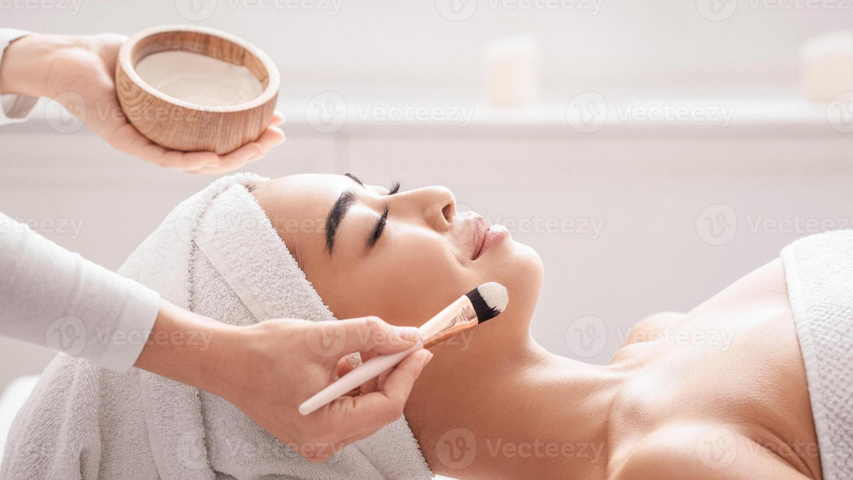 Cosmetologist applying moisturising clay mask on young asian woman face, relaxed beautiful lady with towel on head enjoying skin care treatment procedures at spa salon, side view with copy space photo