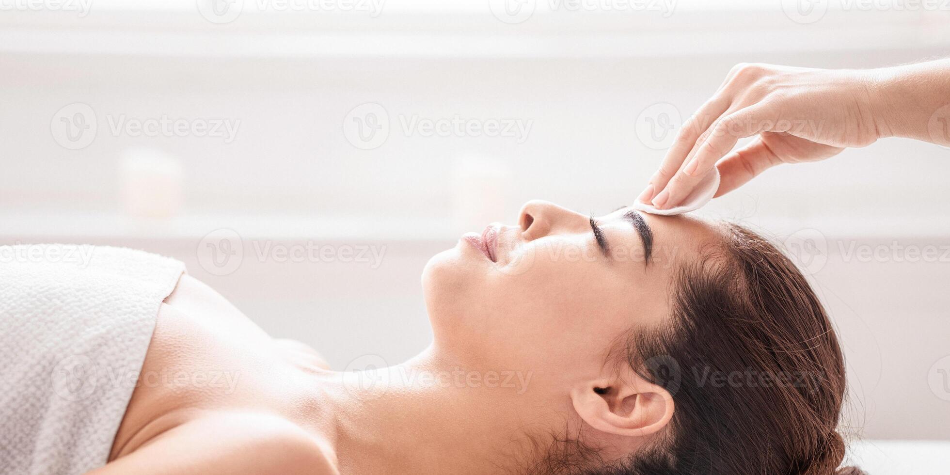 Facial Treatment. Professional Cosmetologist Cleansing Skin Of Young Asian Woman Before Beauty Procedures, Using Cotton Pad, Relaxed Korean Female Lying On Massage Table With Closed Eyes, Side View photo