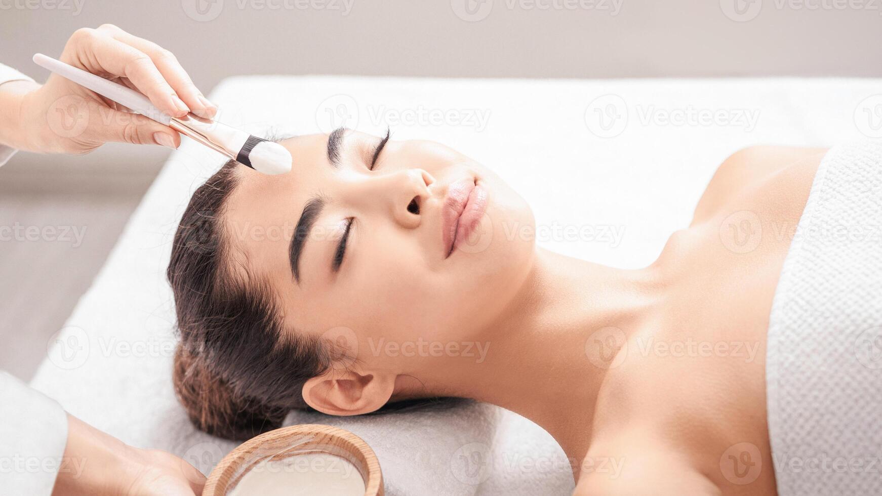 Beauty Treatment. Professional beautician applying clay mask to asian woman at spa salon, young attractive korean female lying on massage table, enjoying face care procedures, closeup shot, free space photo