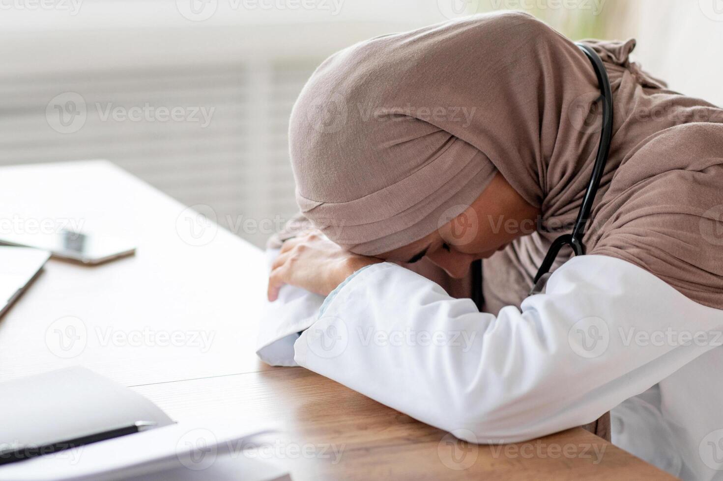 Tired female doctor woman in hijab having nap at workdesk, working with laptop in clinic. Exhausted physician young lady leaning on her hands, needs for break during long working day, copy space photo