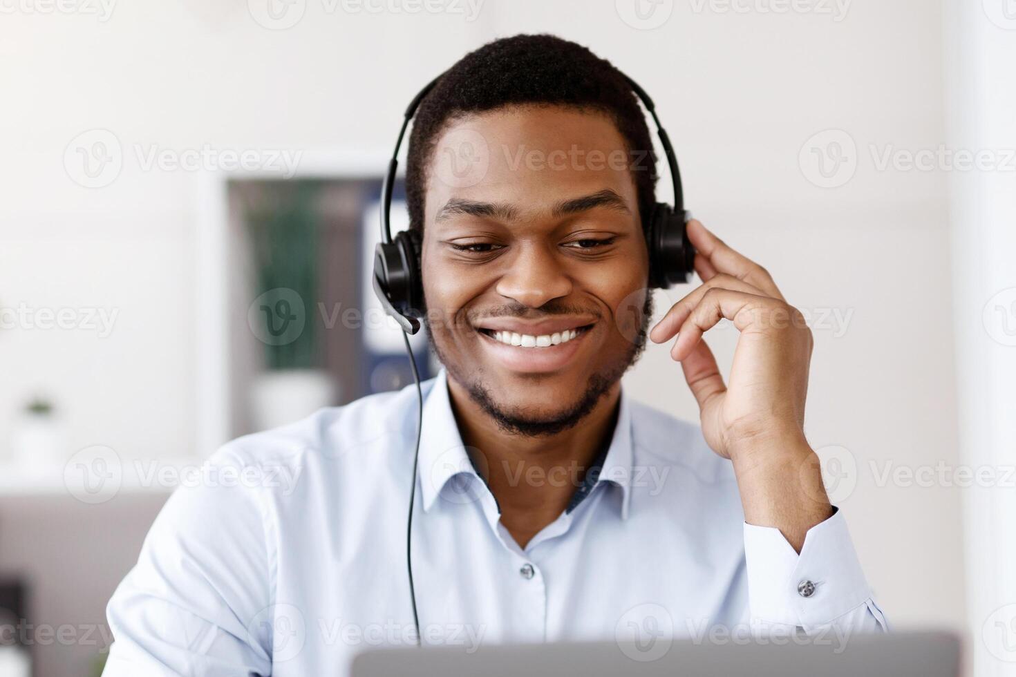 Closeup of black guy worker with headset using laptop, cheerfully talking to client or business partner, smiling at computer screen, african american man having business training, copy space photo