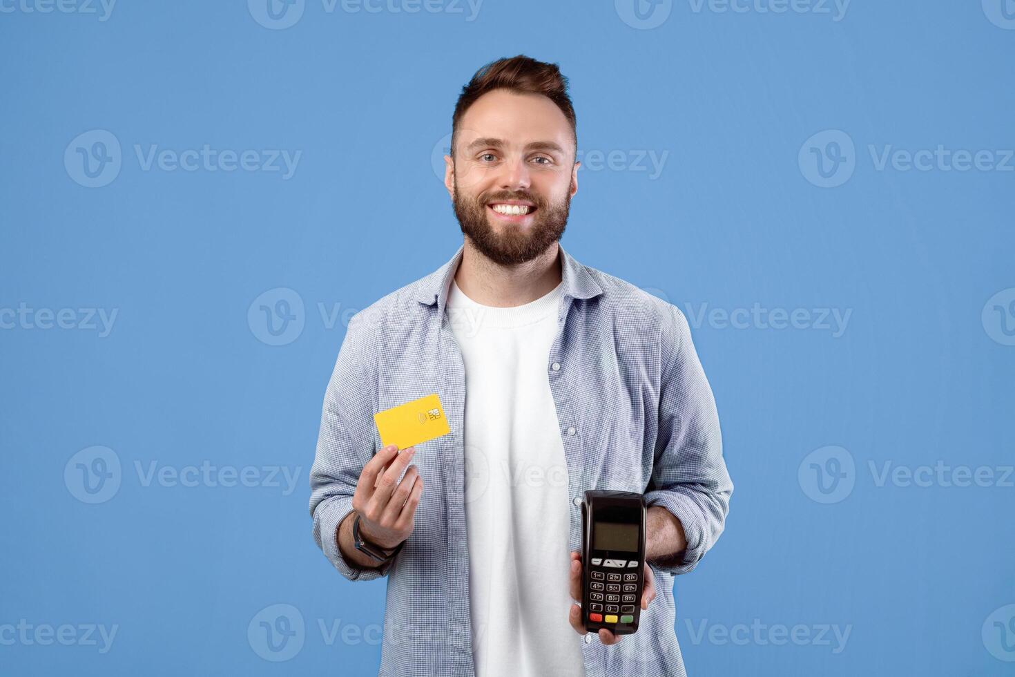 Smiling millennial guy holding payment terminal and credit card, looking at camera on blue studio background. Cool young man using electronic currency, preferring contactless shopping photo