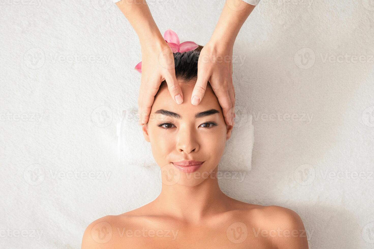 Young asian woman enjoying anti-aging facial massage at spa salon. Female masseur therapist making head and face beauty treatment to relaxed korean lady, top view, cropped image with copy space photo