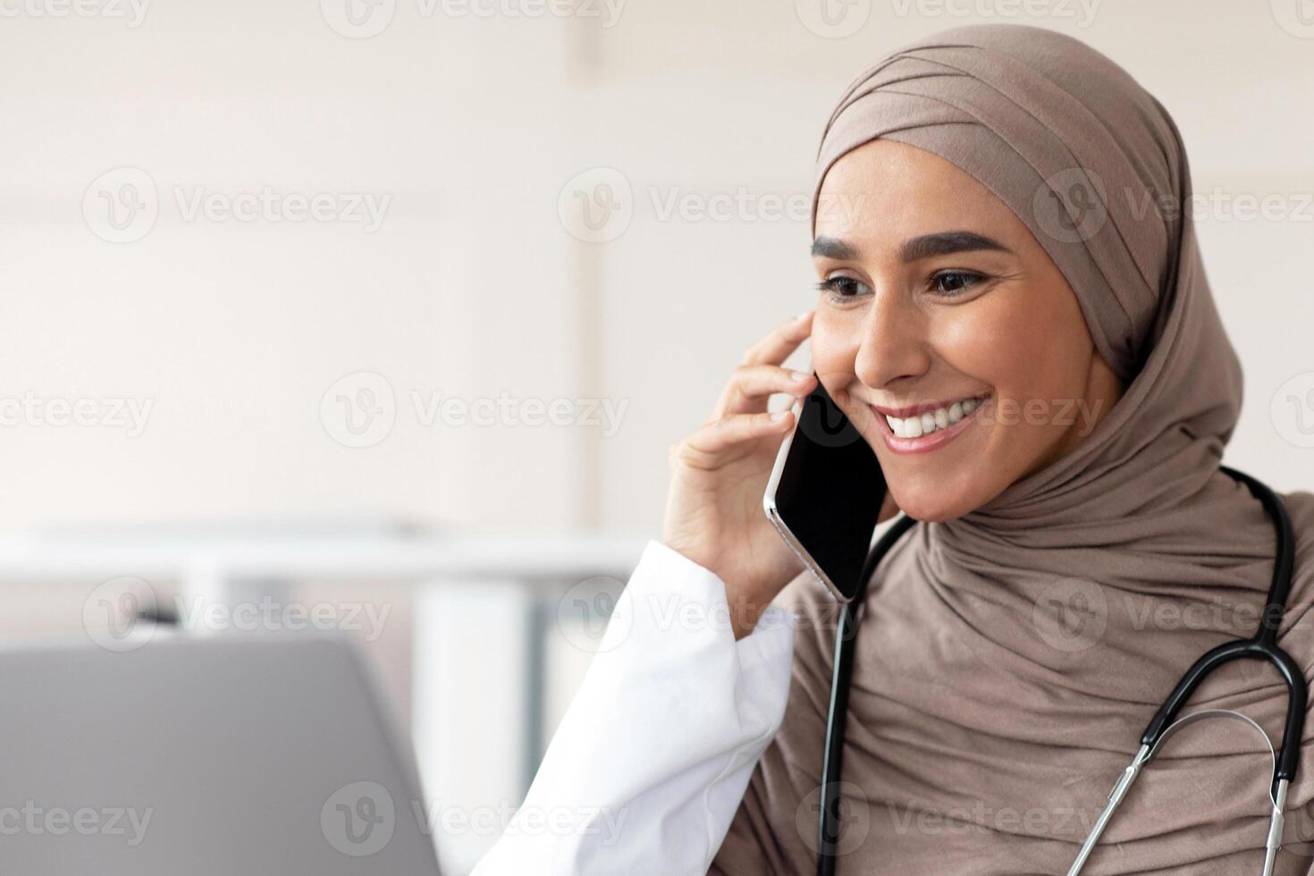 Cheerful muslim lady doctor having conversation on mobile phone with patient, looking at laptop screen, talking to sick client while working at clinic, copy space. Female physician having appointment photo