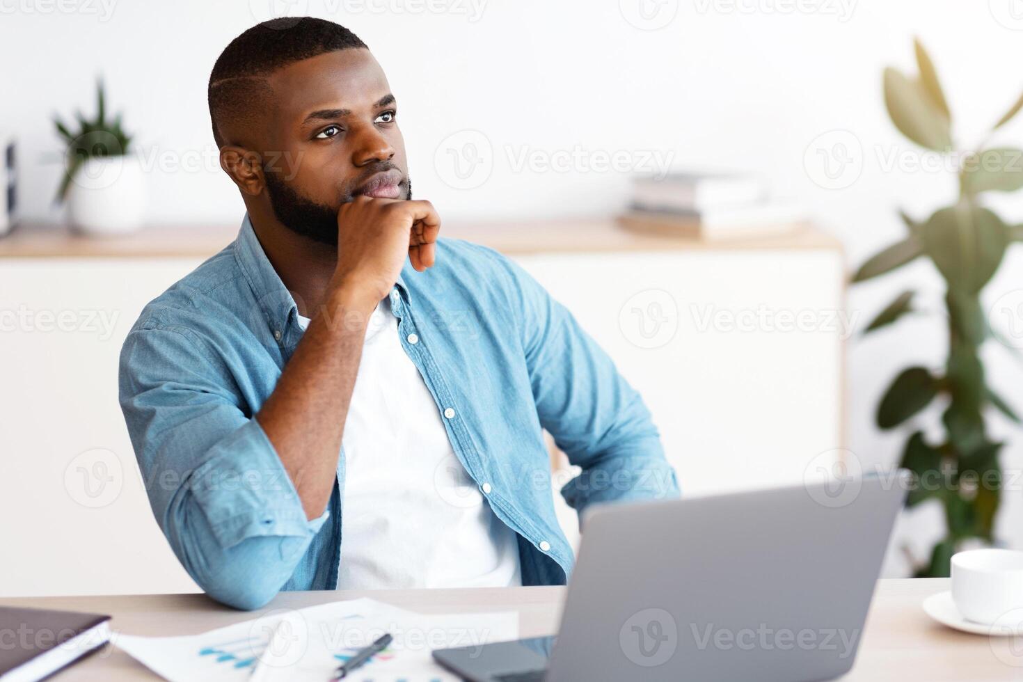 Black Freelancer Guy Sitting At Workplace With Thoughtful Face Expression, Pensive African American Man Looking Away, Waiting For Inspiration While Working With Laptop At Home Office, Free Space photo