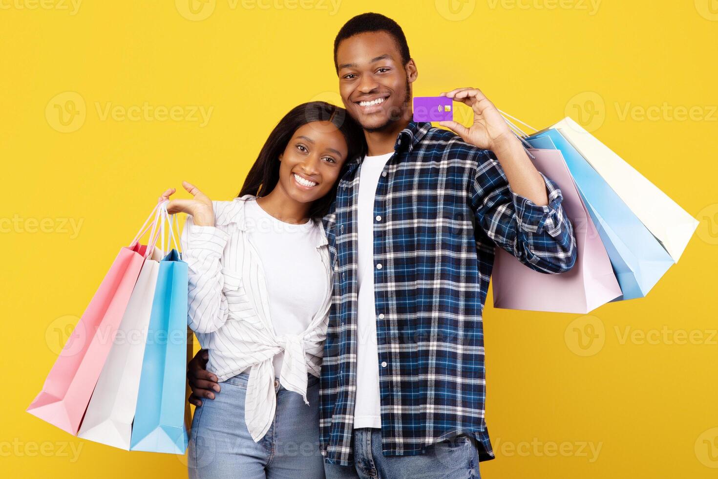 Shopping, free time and seasonal sale together. Cheerful young african american couple in love hold colored bags, hug and show credit card, isolated on yellow background, studio shot, copy space photo