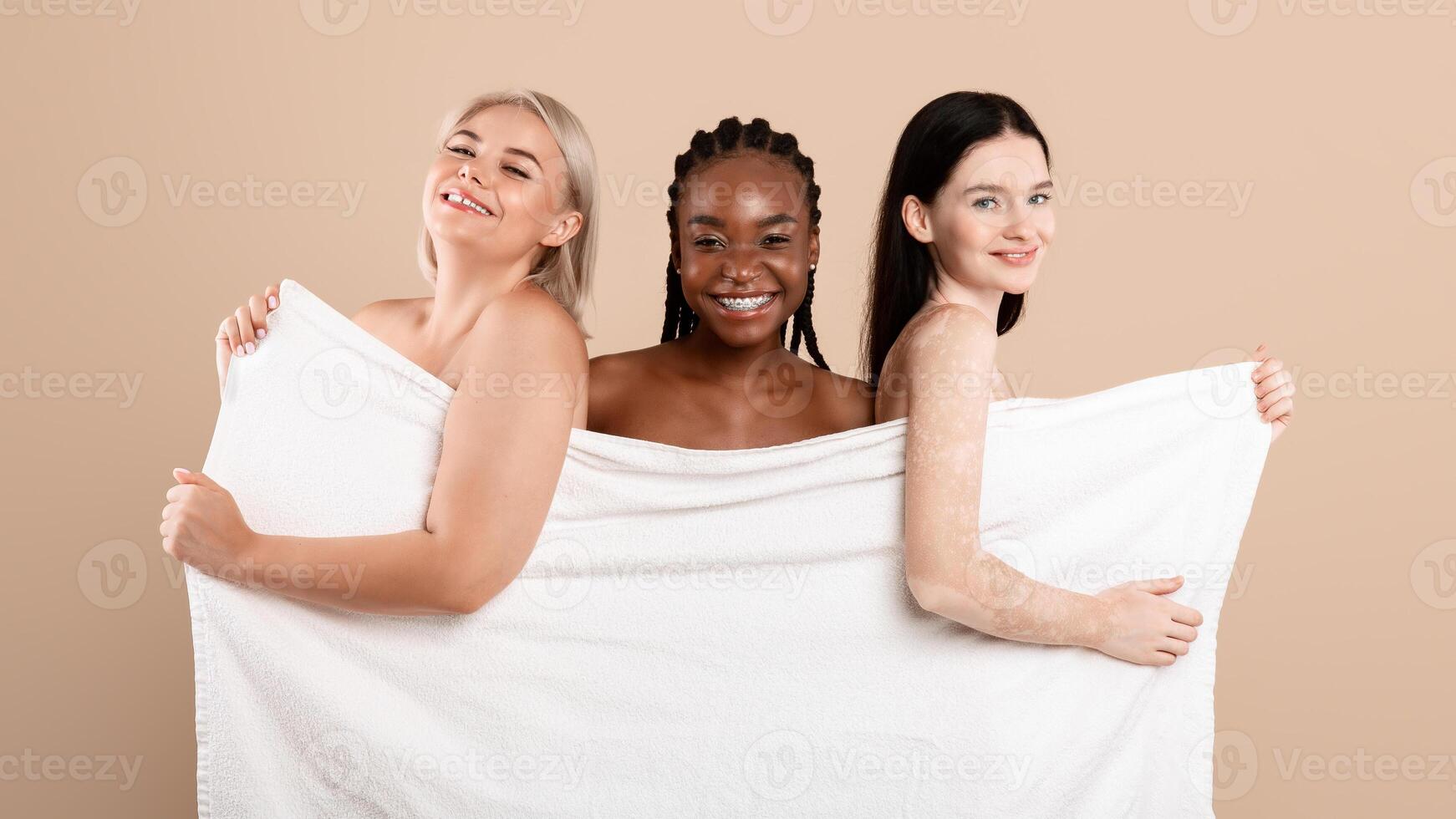 Body Care Concept. Group Of Happy Multiethnic Ladies With Different Skin And Beauty Types Standing Behind Big Bath Towel, Cheerful Multicultural Girlfriends Having Fun Together On Beige Background photo