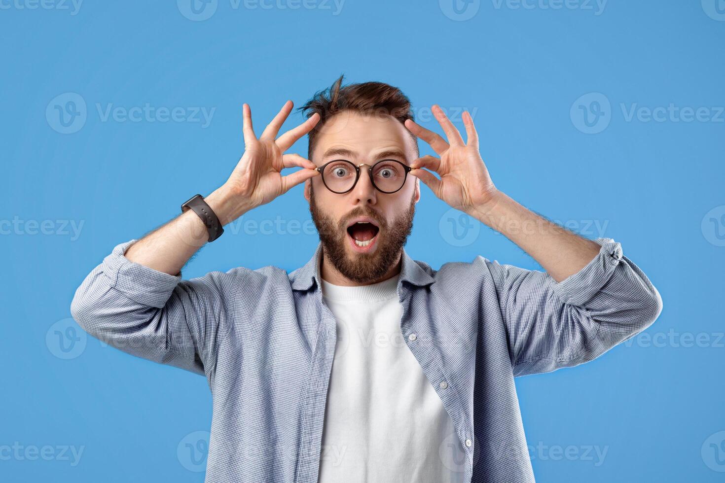 Unbelievable offer. Amazed Caucasian guy in casual outfit and glasses opening mouth in surprise on blue studio background. Attractive bearded man feeling shocked and stunned, saying OMG photo