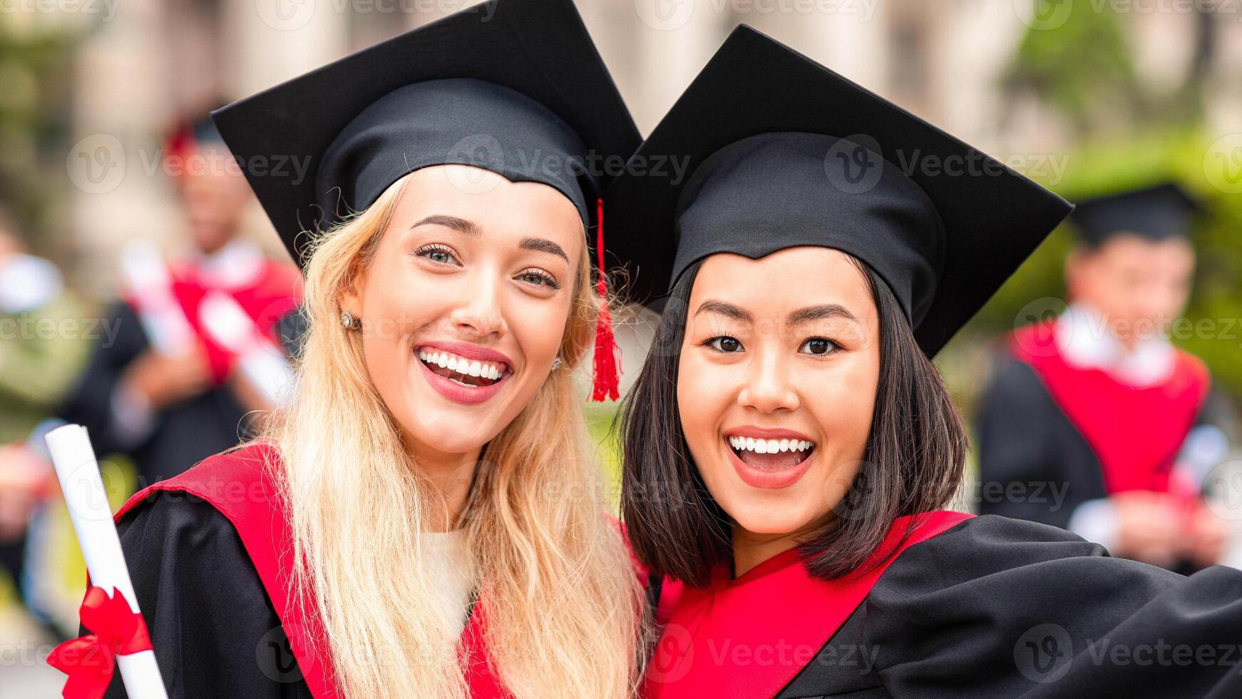 Two cheerful multiracial young ladies girlfriends in graduation clothes taking selfie, happy female students showing diplomas and smiling at camera, closeup portrait over university campus photo