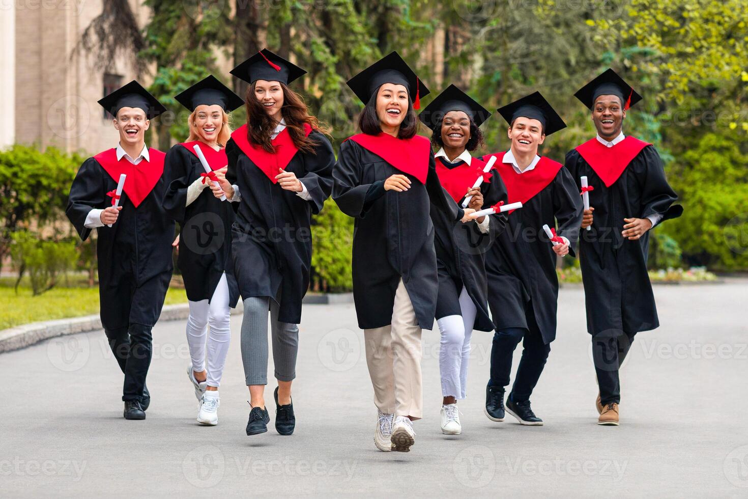 Cheerful multiracial students guys and young ladies in graduation costumes walking by university campus, holding diplomas and smiling. College, university graduation, education concept photo