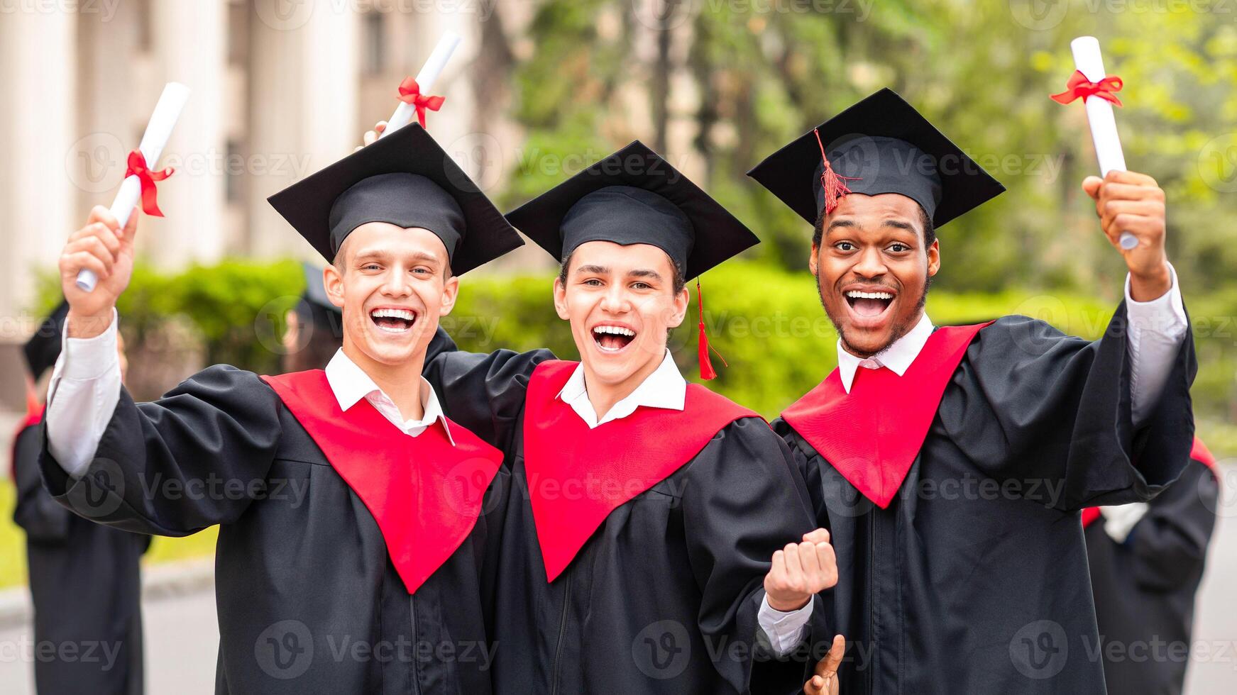 Positive guys students posing together next to university, wearing graduation costumes and showing diplomas. Cheerful multiracial group of male friends celebrating graduation, portrait photo