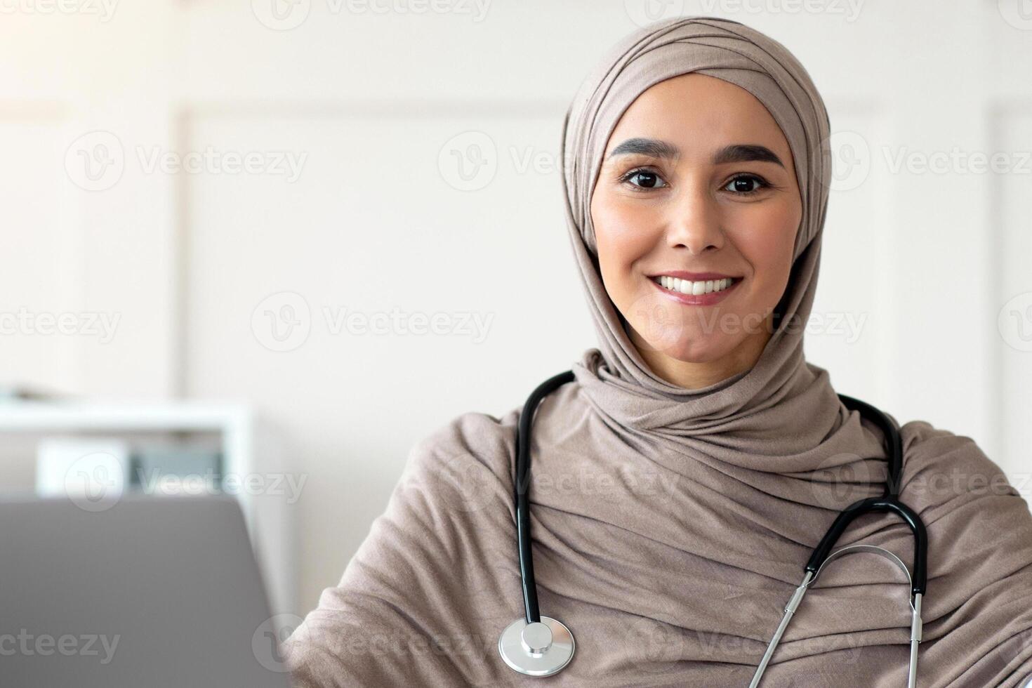 Beautiful muslim woman in hijab doctor with stethoscope posing in hospital, sitting at worktable with laptop, medical folder, smiling at camera, copy space. Doctor's office, cabinet interior concept photo