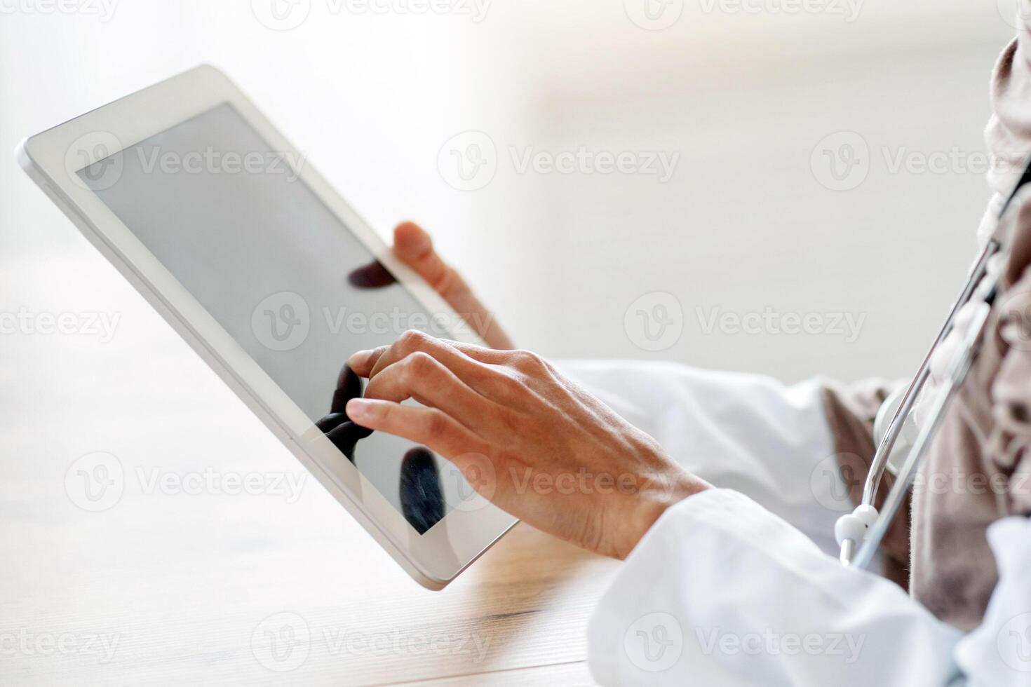 Cropped of female doctor in hijab using gadgets at workplace, holding digital tablet. Unrecognizable therapist muslim woman having online conversation with her patients, using pad, side view photo