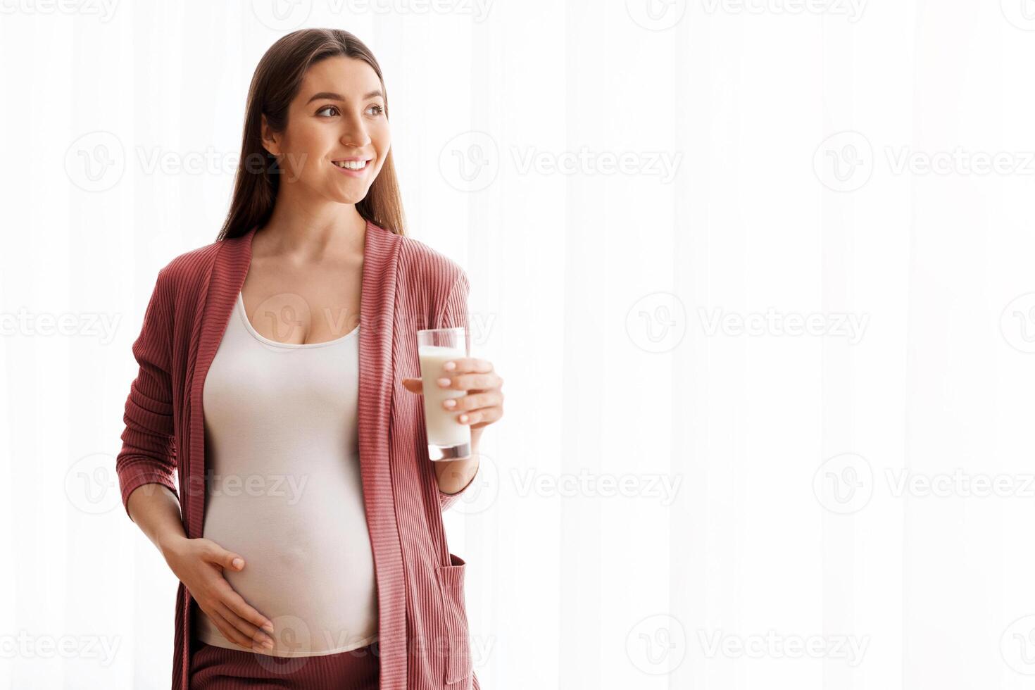 Happy Pregnant Lady Enjoying Healthy Calcium Drink, Holding Glass With Milk While Standing Near Window At Home, Young Smiling Expectant Lady Tenderly Embracing Belly And Looking Away, Copy Space photo