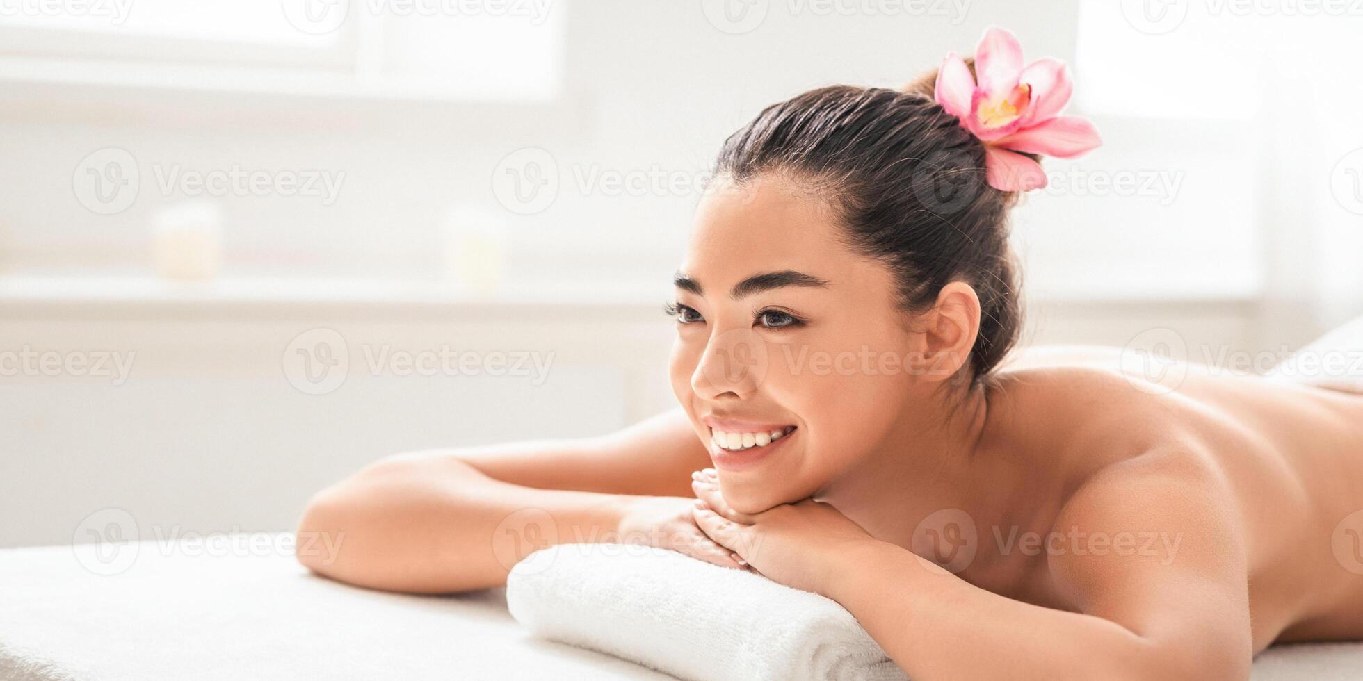 Beautiful Young Asian Woman Enjoying Wellness Day In Spa Salon, Smiling Korean Lady With Exotic Flower In Hair Lying On Massage Table, Waiting For Beauty Treatment, Closeup Shot With Copy Space photo