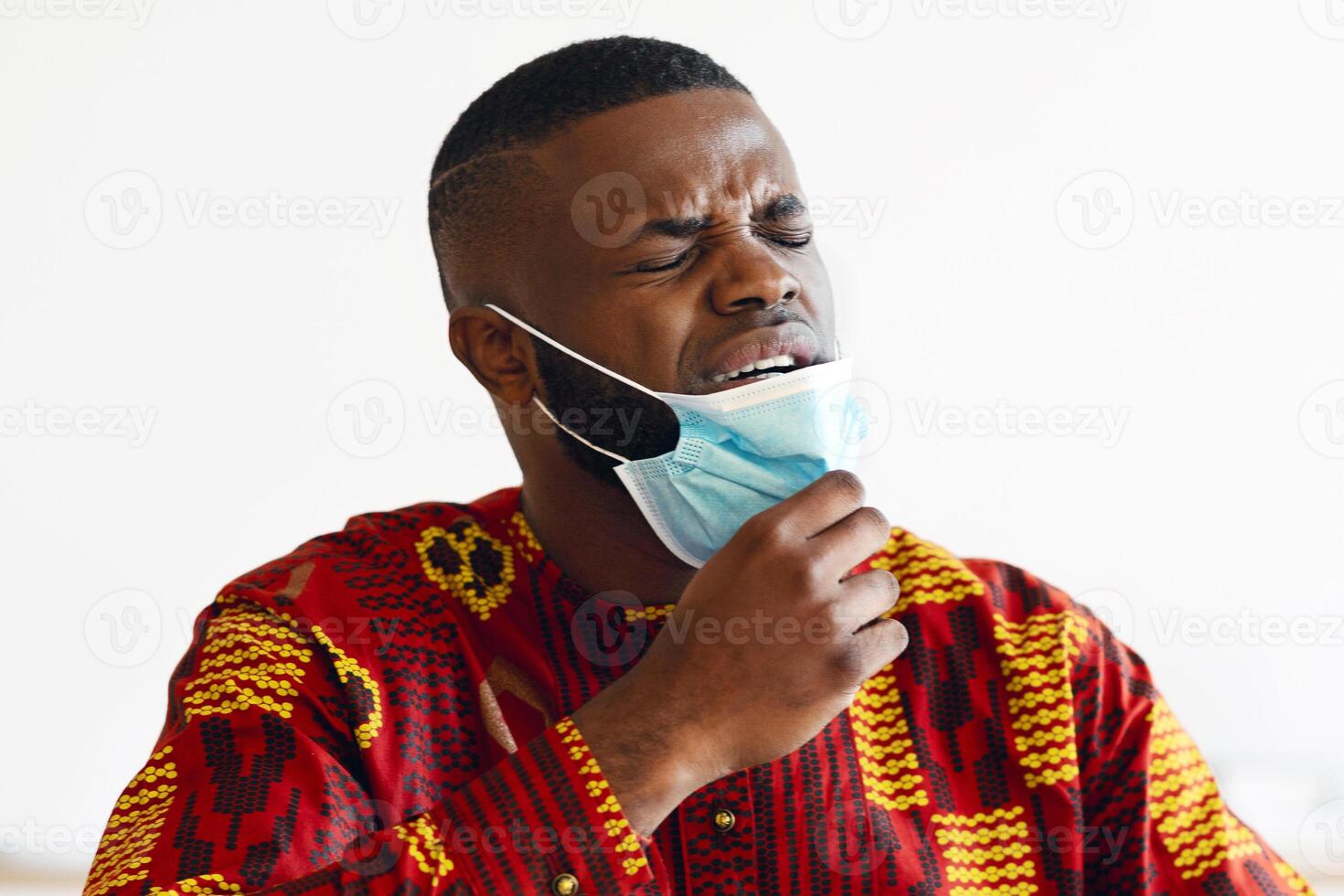 Young African Man Taking Off Medical Mask At Work, Can Not Breathe, Black Guy In Traditional Clothes Suffering From Allergy At Workplace, Sneezing, Having Symptoms Of Coronavirus, Copy Space photo