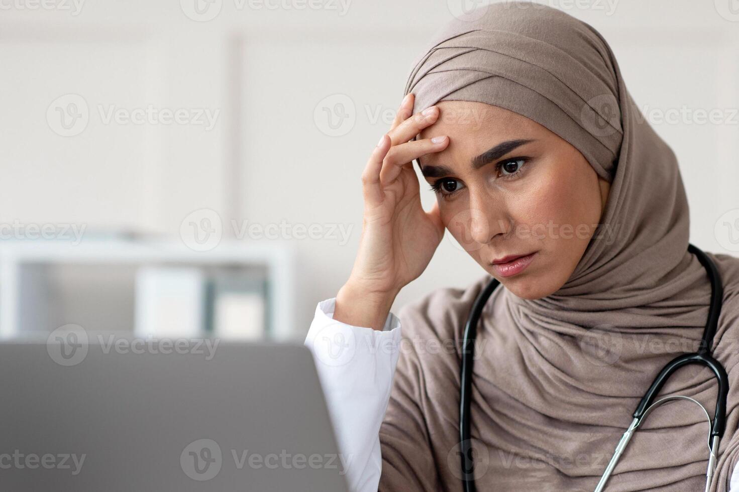 Tired female doctor beautiful woman in hijab having headache while working with laptop, typing on computer keyboard and touching her forehead, closeup, copy space, clinic interior photo