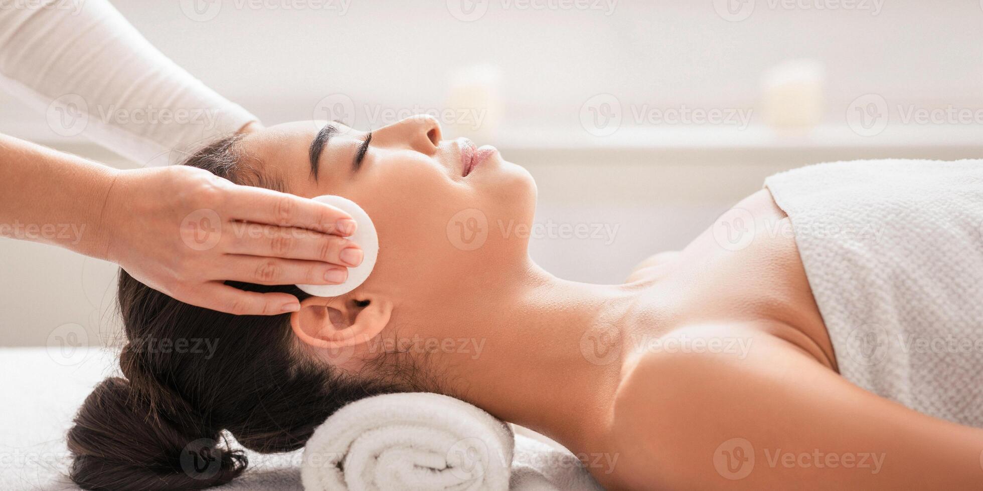 Professional Cosmetologist Cleansing Skin Of Young Asian Woman During Facial Beauty Treatments In Spa, Relaxed Korean Lady Enjoying Wellness Day At Beauty Parlor, Lying In Massage Table, Side View photo