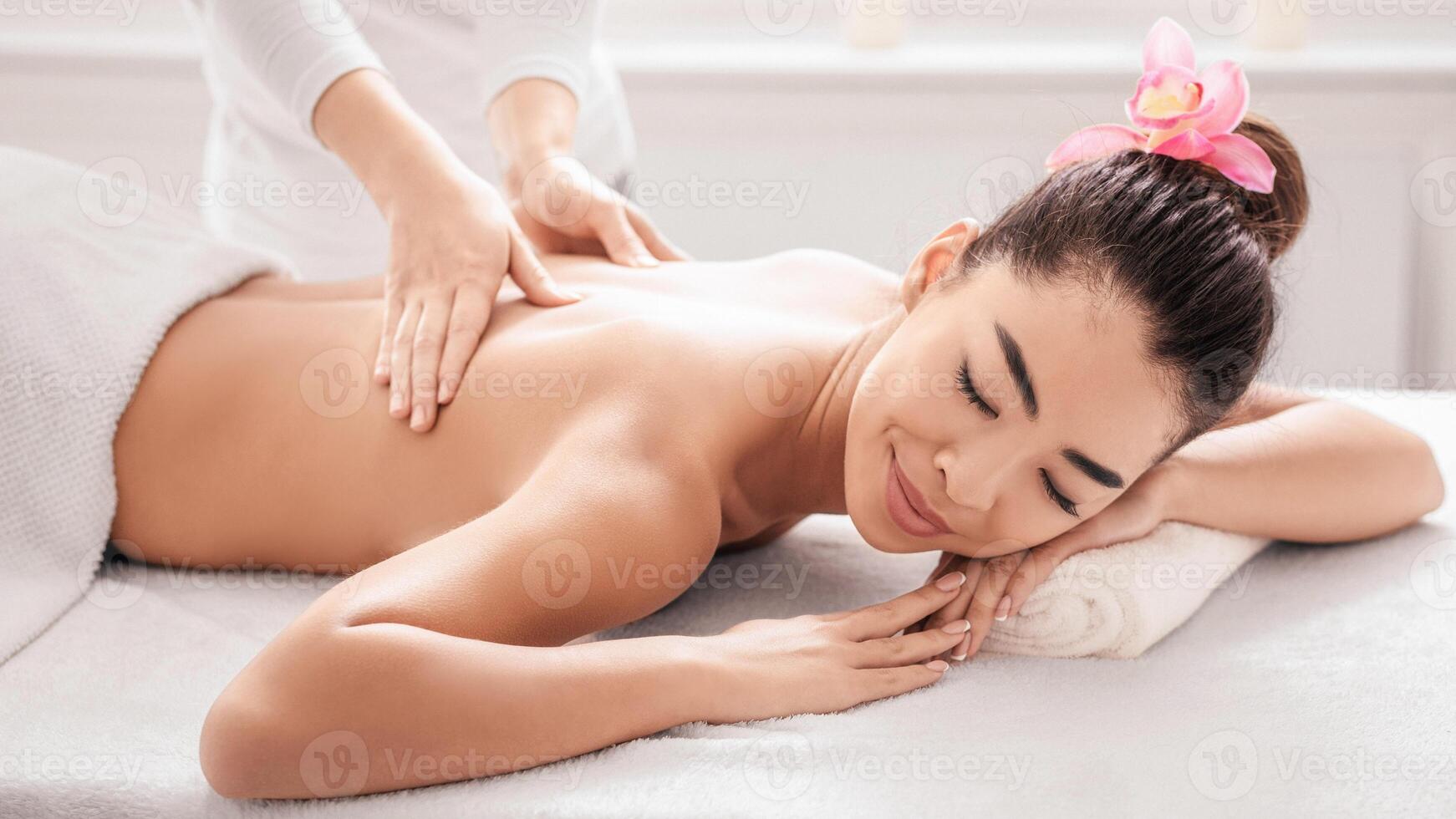 Young Asian Woman Enjoying Back Massage In Spa Salon, Getting Relaxing Beauty Treatment From Professional Masseur, Calm Korean Lady Lying On Table With Closed Eyes, Side View, Closeup Shot photo