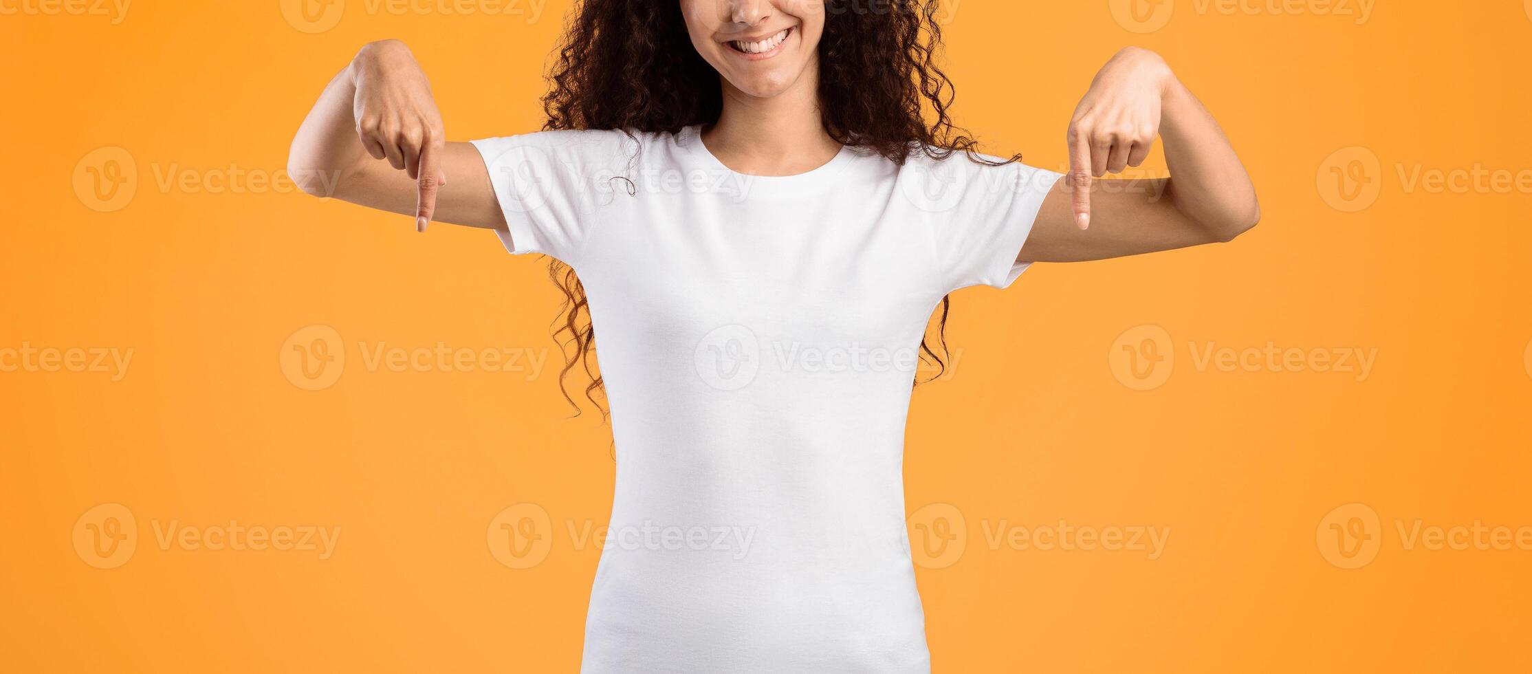 Happy Curly Woman Pointing Fingers Down Posing Smiling To Camera Standing In Studio Over Yellow Background, Wearing White T-Shirt. Look There Advertisement Banner. Panorama photo