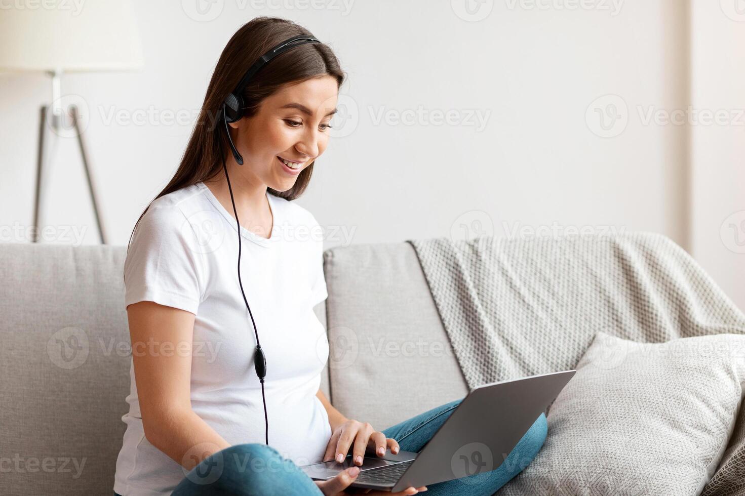 Cheerful Pregnant Woman In Headset Using Laptop At Home, Studying Or Working Online. Smiling Expectant Mother Watching Webinar Or Having Training In Internet, Enjoying Distance Learning And Remote Job photo