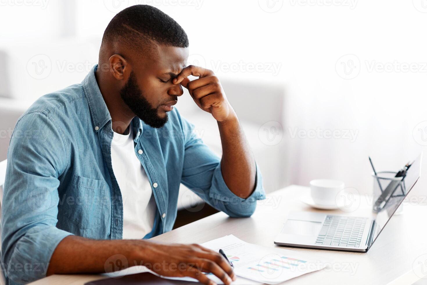 Migraine Concept. Overworked African American Man Suffering From Headache At Workplace, Stressed Black Freelancer Guy Sitting At Desk With Laptop And Documents And Touching Nose Bridge, Feeling Unwell photo