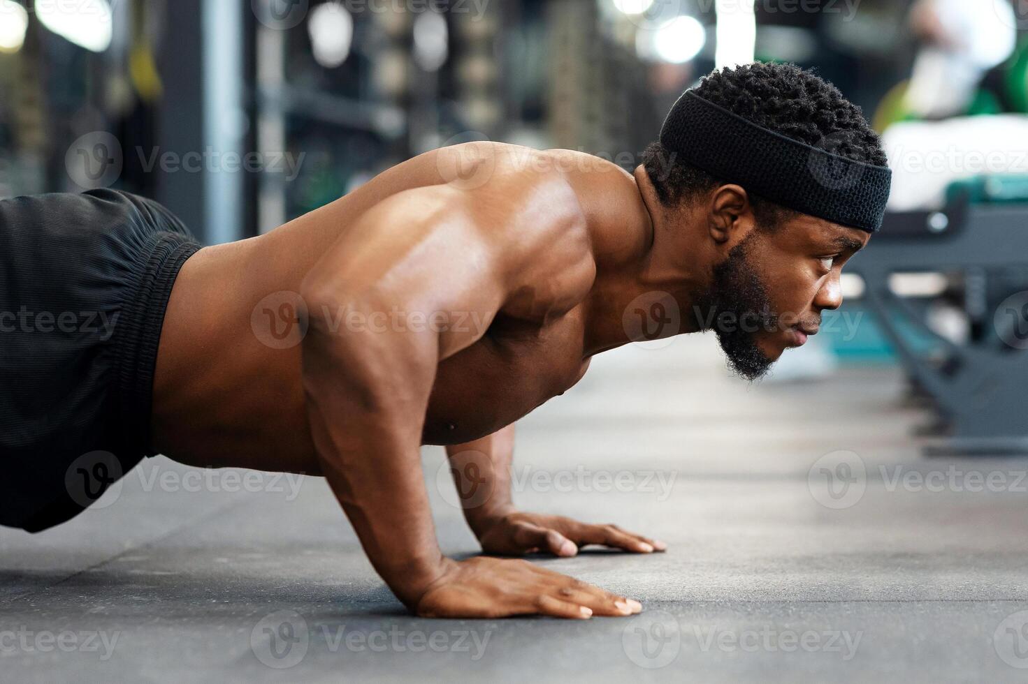 Push-ups, press-ups workout. Muscular african american shirtless guy sportsman building up muscles, black man training on floor at modern gym, doing push-ups and looking at copy space, panorama photo