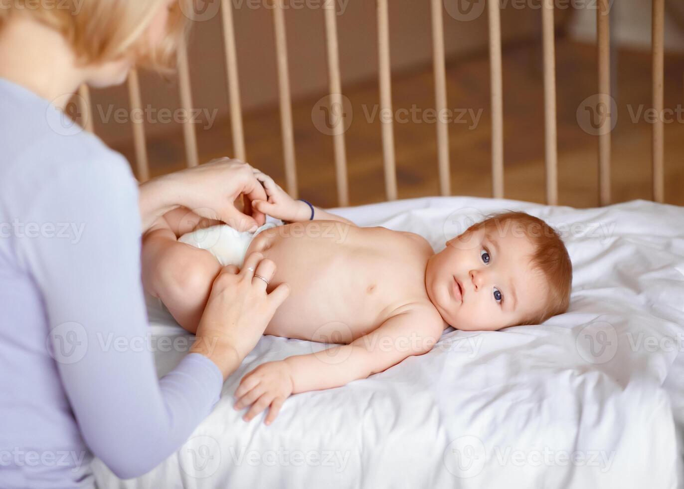Beautiful baby boy looking at camera while his mother changing nappy, baby girl woke up after nap, laying in wooden baby crib. Mom changing diaper for her little kid, cropped, copy space photo