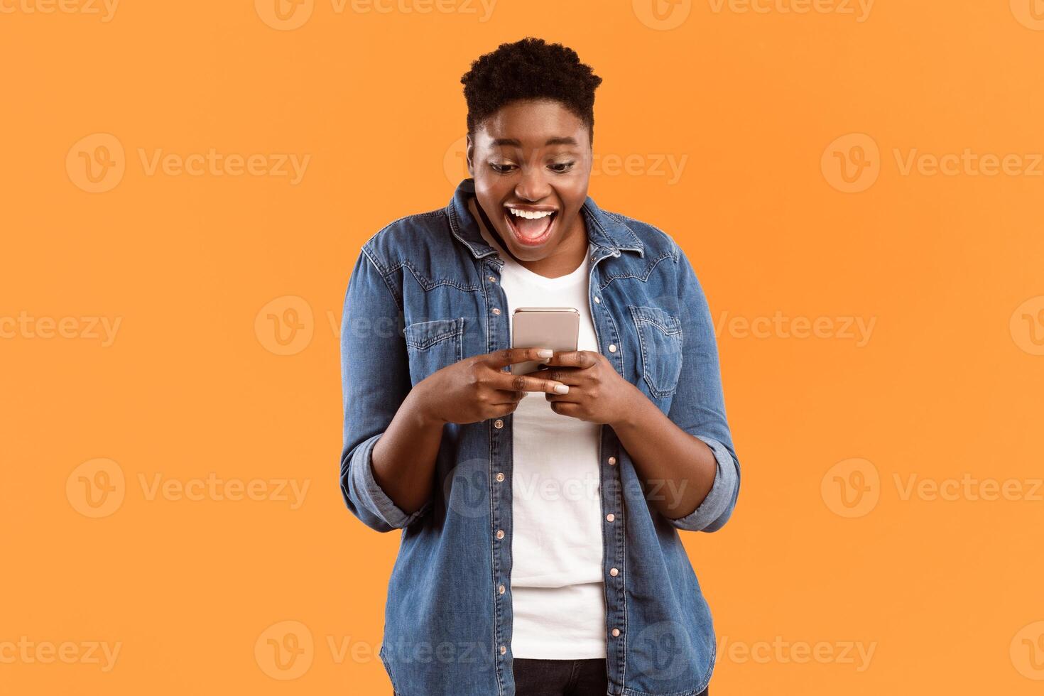 Excited African American Lady Using Cellphone Texting In Social Media Or Playing Mobile Game Standing Over Yellow Studio Background. Great Mobile Application For Your Smartphone Concept photo