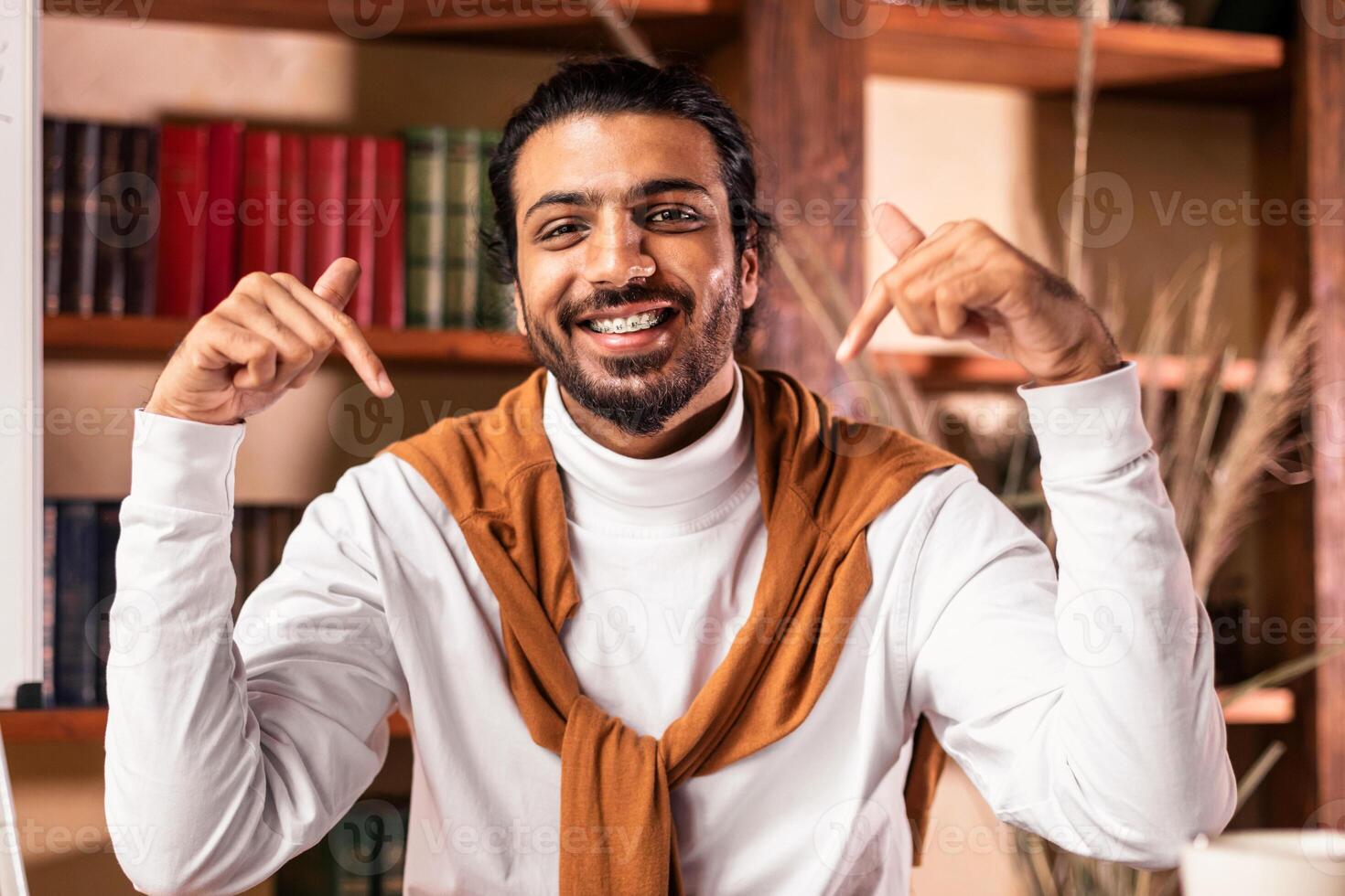 Choose online education. Indian teacher pointing at himself and laptop recommending virtual e-learning sitting near blackboard indoors, smiling to camera. Selective focus photo