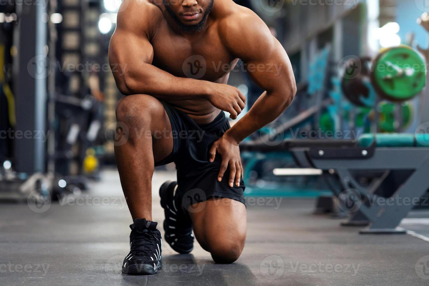 Tired african american shirtless bodybuilder having break after workout at modern gym. Serious black guy sportsman resting after exercising, leaning on one knee, copy space photo