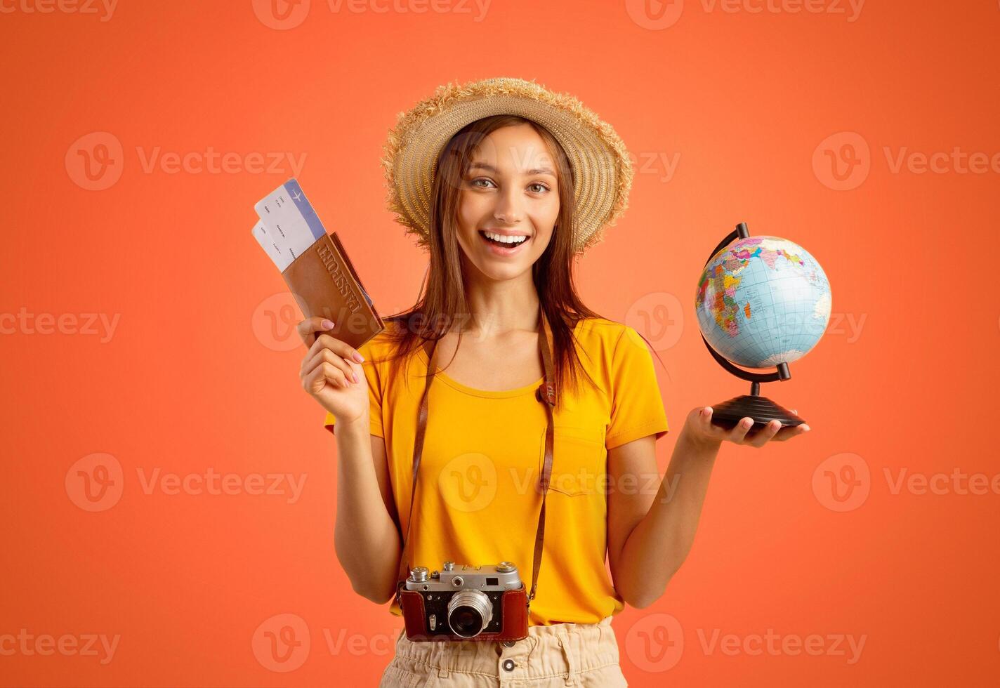 Cheerful young woman in summer hat with passport, tickets, globe and camera over yellow studio background, going vacation, copy space. Female traveller travelling abroad, having exciting trip photo