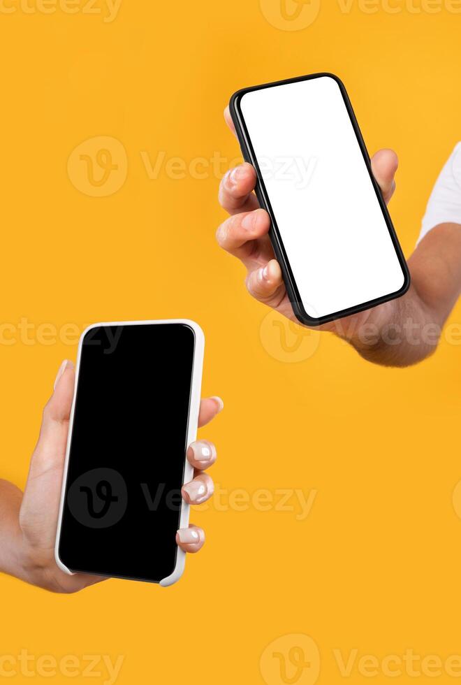 Closeup of female and male hands holding 2 smartphones with white and black empty screen, showing devices close to camera. Two gadgets with blank space, mock up, selective focus, vertical shot photo