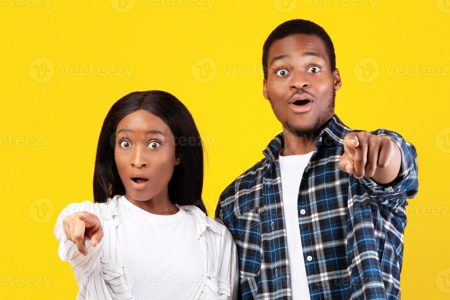 Wow, what is this. This is unimaginable and amazing. Shocked teen african american students man and lady in casual with wide open eyes pointing fingers at camera, isolated on yellow background photo