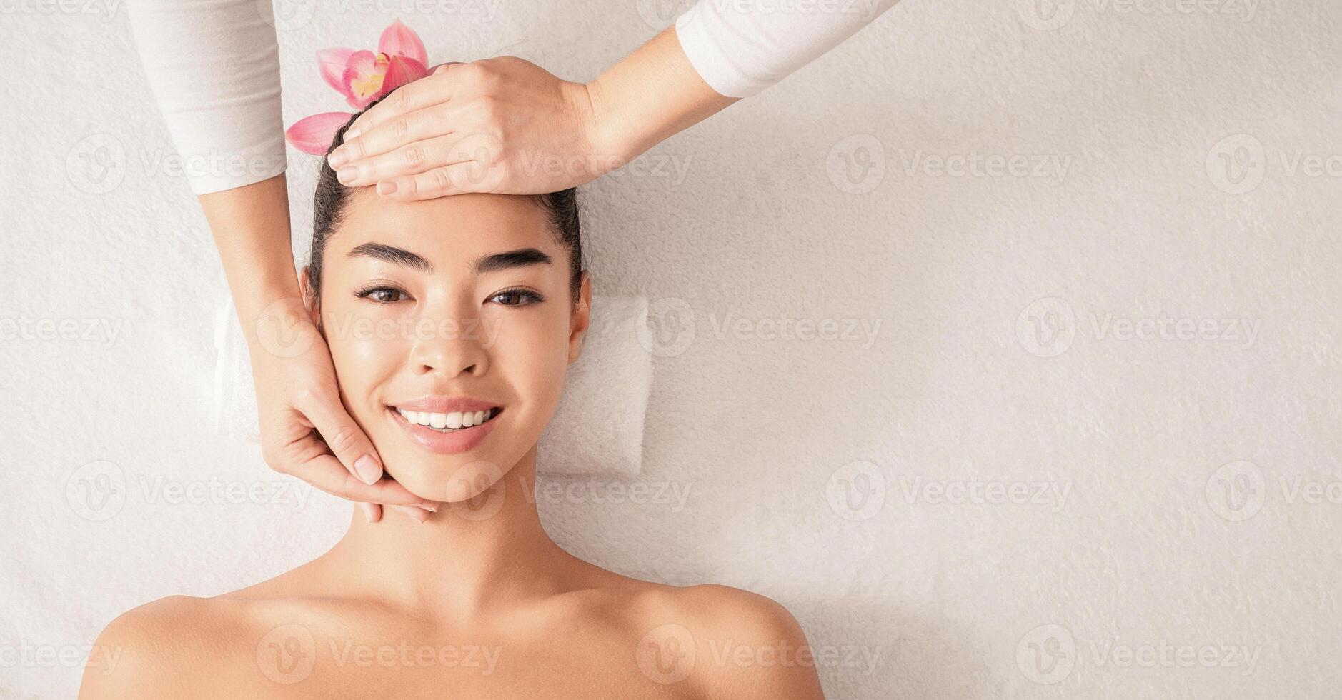 Relaxing Massage. Portrait Of Beautiful Asian Woman Enjoying Facial Beauty Treatments At Spa Salon, Smiling Young Korean Female With Flower In Hair Having Wellness Day, Top View With Copy Space photo