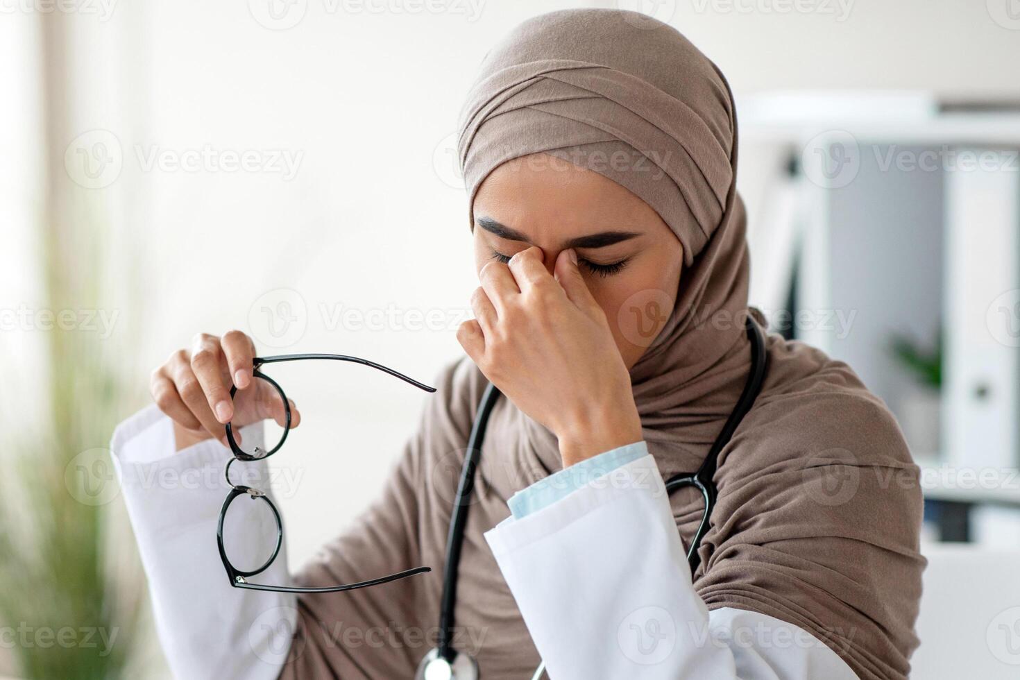 Muslim doctor young woman in hijab with stethoscope having eyesight problem while working with laptop at clinic, holding glasses and rubbing her nose bridge with closed eyes, copy space photo