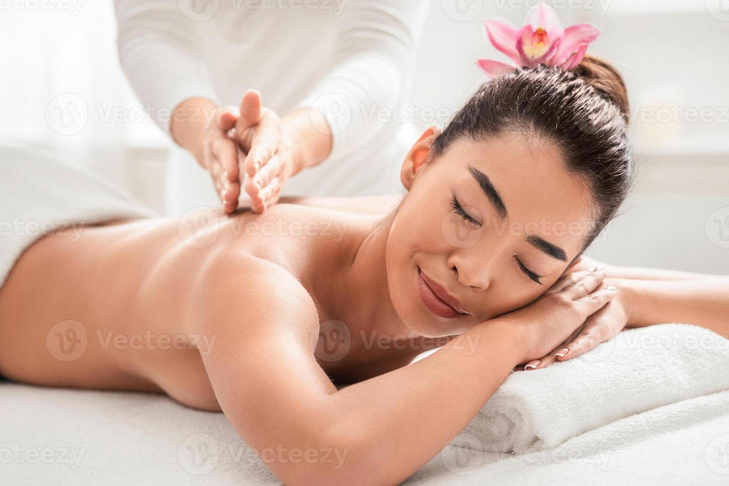 Young Korean Lady Having Relaxing Back Massage Session In Spa Salon, Beautiful Asian Woman With Flower In Hair Getting Treatment With Masseur, Lying With Closed Eyes, Enjoying Wellness Day, Closeup photo