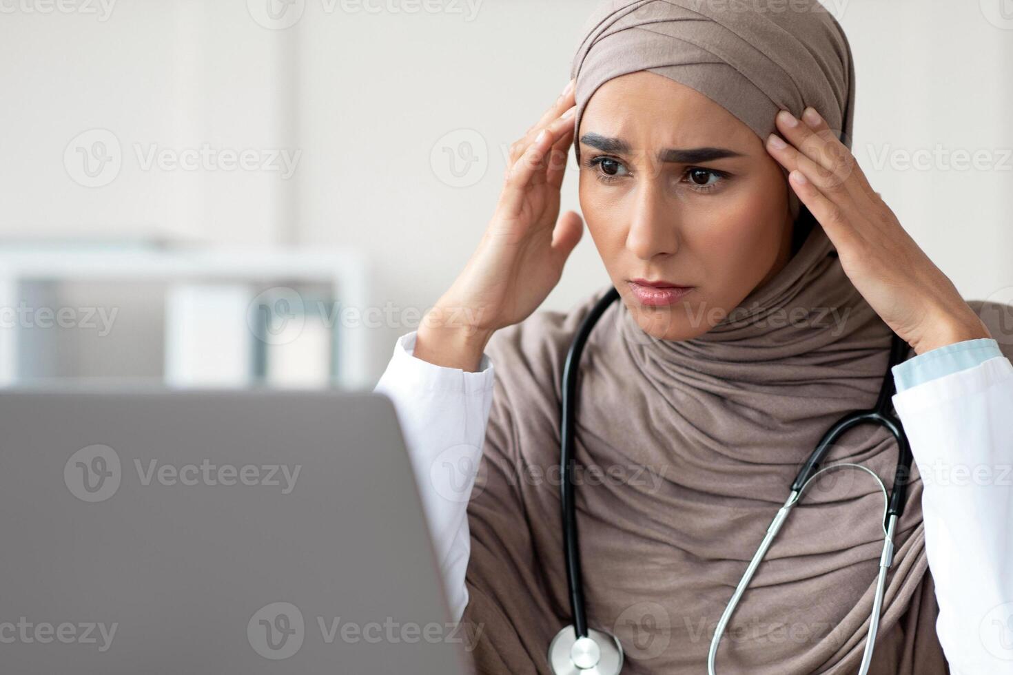 Worried woman doctor beautiful woman in hijab having headache while working with laptop, looking at screen and touching her temples, having difficulties at job, closeup, copy space, clinic interior photo