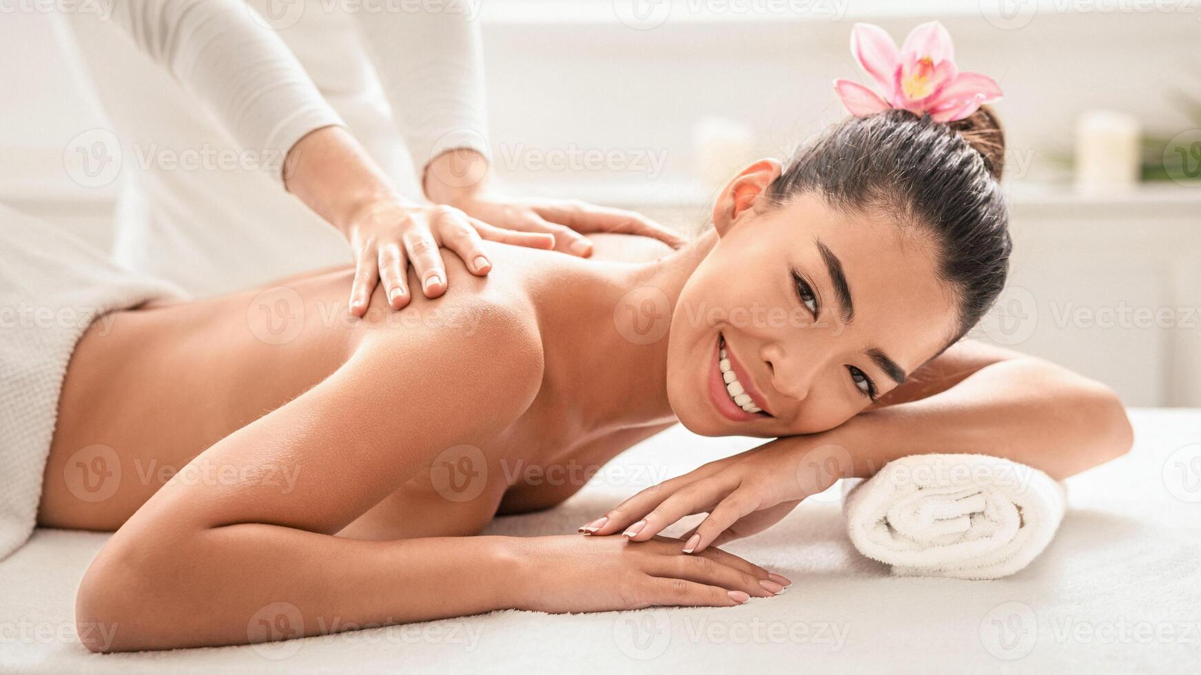 Wellness Concept. Beautiful asian lady enjoying back massage in spa center, relaxed naked young korean female lying on table with rolled towel under head, smiling at camera, closeup shot photo