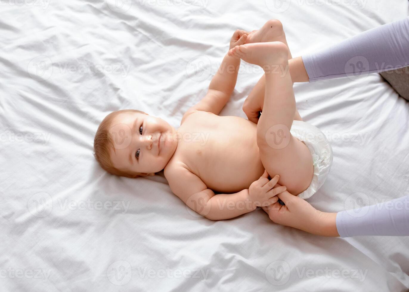 Unrecognizable mother changing her smiling little baby's diaper on white, top view. Cute toddler blonde boy having fun while his mom dressing him up, playing with his feet, closeup, empty space. photo