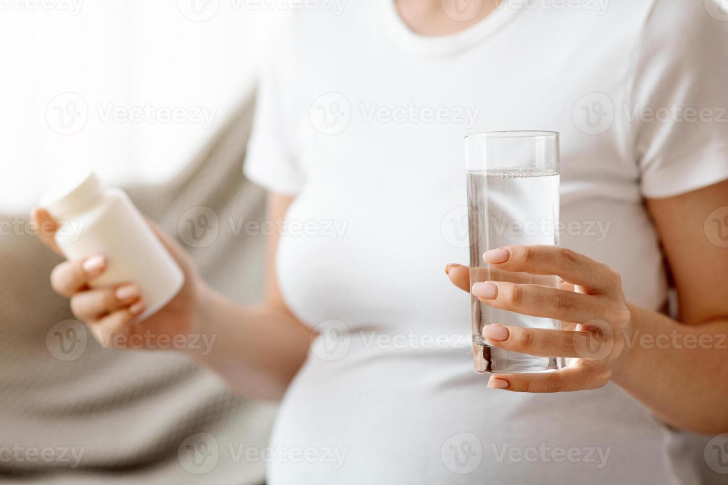 Cropped Image Of Pregnant Woman Holding Jar With Prenatal Vitamins And Glass Of Water While Sitting On Couch, Unrecognizable Expectant Lady Using Supplements For Healthy Pregnancy, Closeup Shot photo