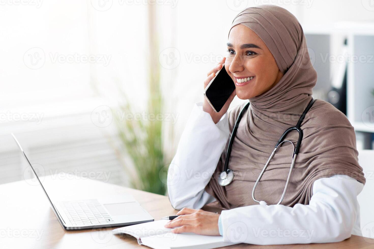 Happy female doctor young muslim woman in hijab having phone call while working at clinic, sitting at workdesk in front of laptop, taking notes at notebook, looking at copy space photo