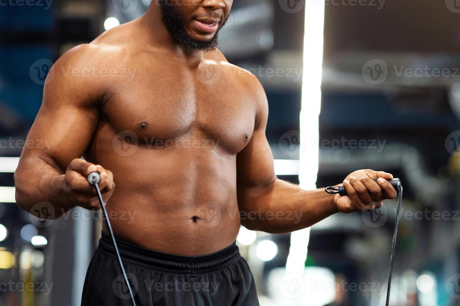 Hot african american young man bodybuilder warming up before working out in gym, closeup. Shirtless muscular black guy jumping with skipping, looking at free space while making cardio photo