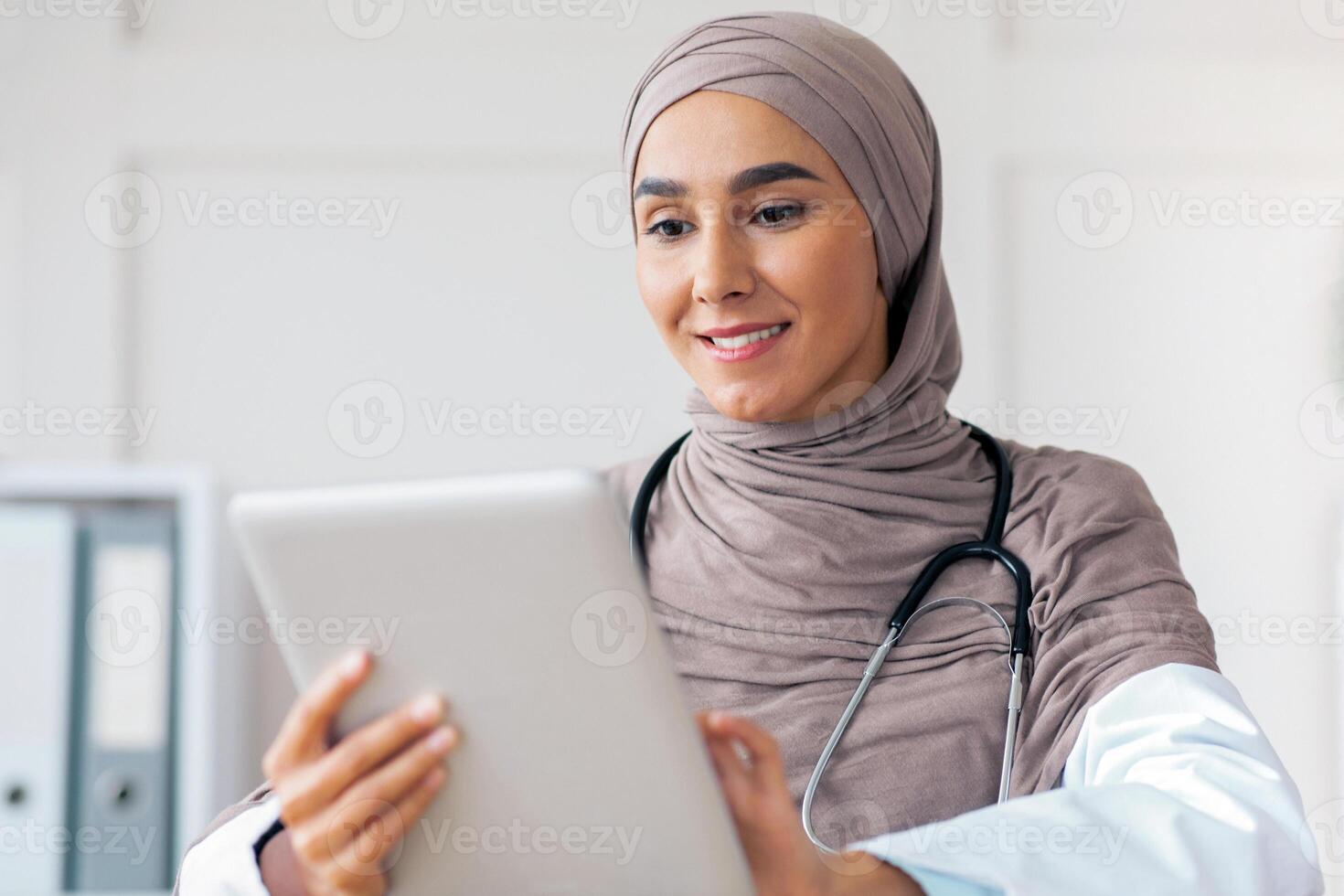 Online appointment with doctor concept. Attractive muslim woman doctor working in clinic, sitting at workdesk and using digital tablet, making online appointment with patient or training photo