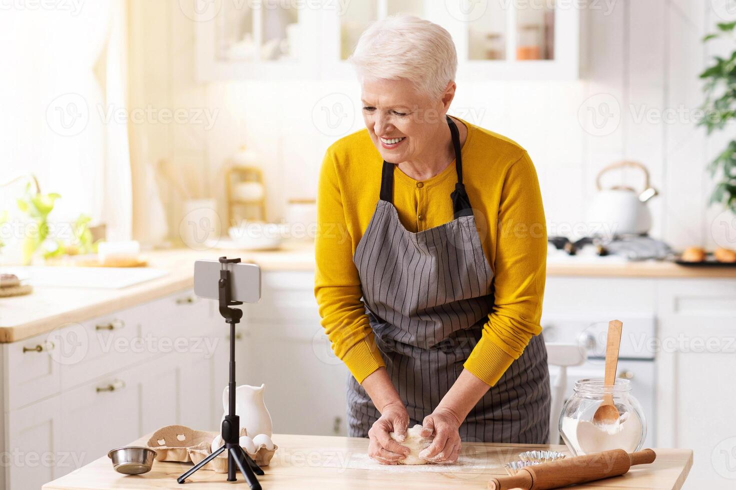Positive old lady food blogger making dough in front of camera, kitchen interior, copy space. Cheerful elderly lady making pastry and shooting videoblog, using mobile phone on tripod photo
