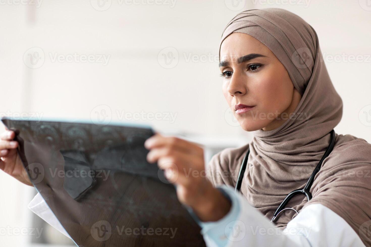 Female doctor muslim woman in headscarf looking at CT scan, working at clinic, empty space. Physician, oncologist, neurologist lady in hijab holding result of tomography, collecting anamnesis photo