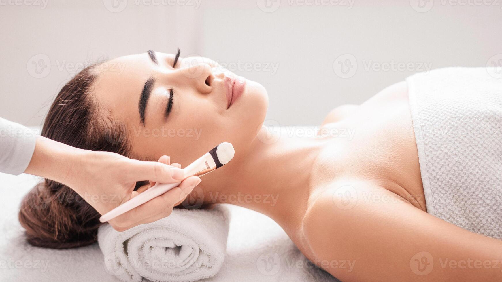 Skin Treatments. Young Korean Lady Enjoying Beauty Procedures In Spa Salon, Lying On Massage Table With Closed Eyes. Professional Beautician Applying Clay Mask To Attractive Asian Woman, Side View photo