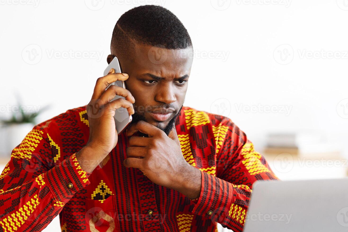 Portrait Of Busy Black Businessman In Tribal Shirt Talking On Cellphone And Working On Laptop, African Entrepreneur In Traditional Clothes Looking At Computer Screen And Having Phone Conversation photo