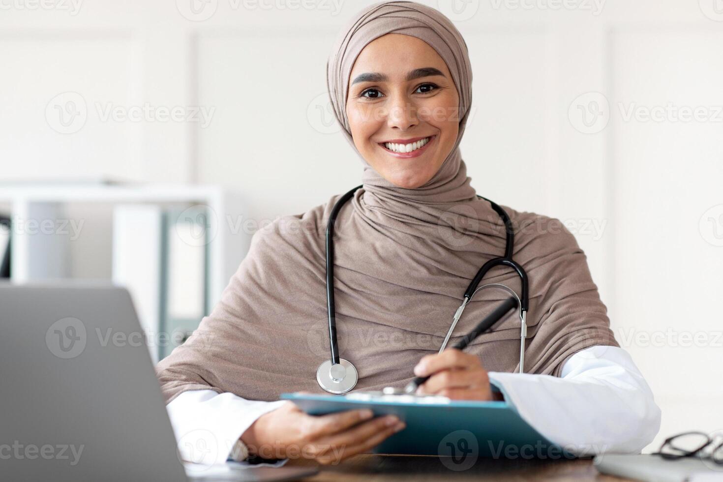 Beautiful muslim woman doctor writing in medical chart and smiling at camera, wearing hijab, sitting at workdesk with modern laptop in her cabinet in clinic or hospital, taking anamnesis, copy space photo