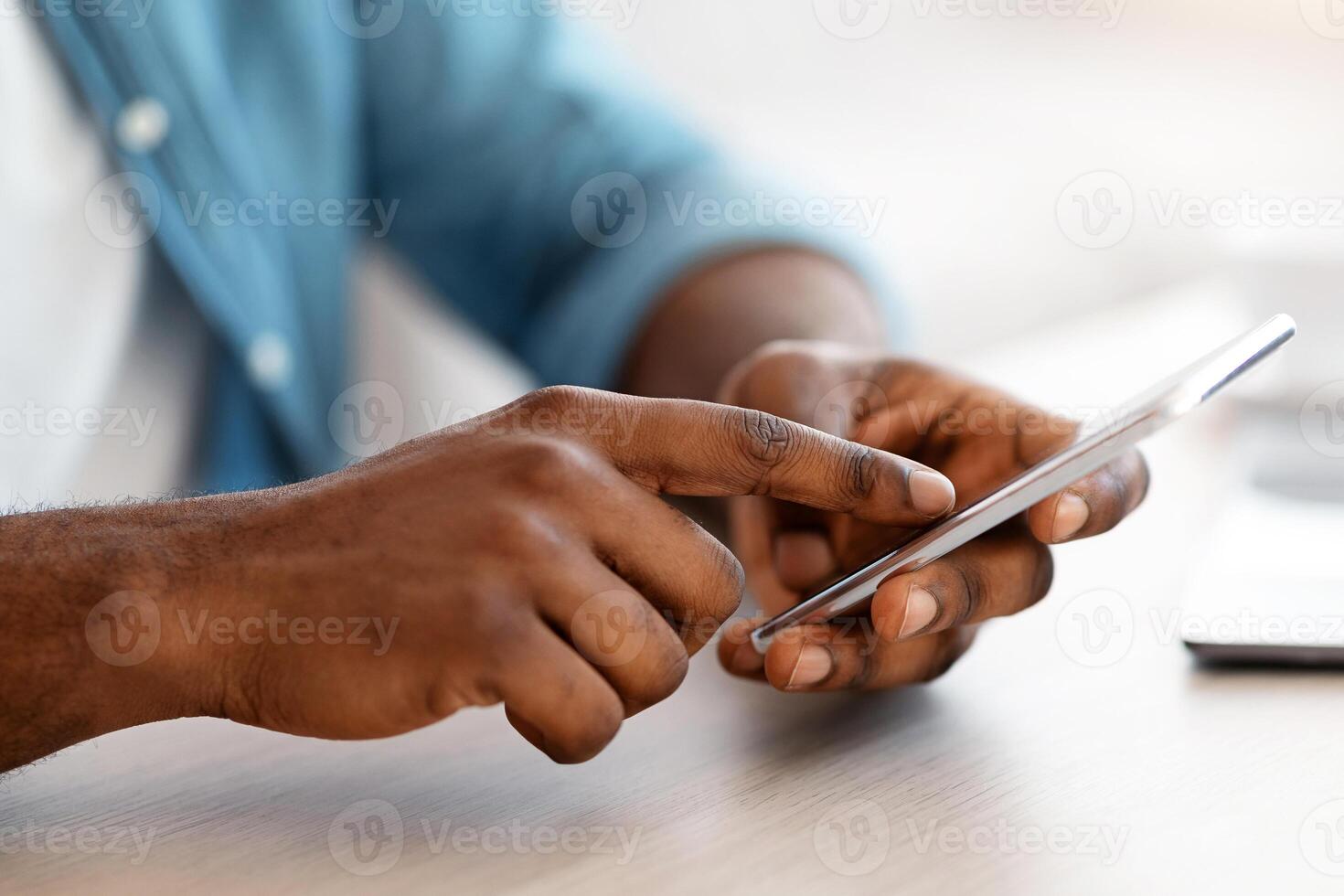 Cropped Image Of Unrecognizable Black Man Using Smartphone While Sitting At Desk In Office, African American Guy Holding Cellphone Browsing New Mobile App Or Shopping Online, Closeup Shot photo